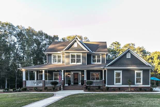 Front view of a large two story blue gray house with wood and vinyl siding with an expansive porch, sidewalk and manicured lawn.